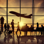 Nella foto è riprodotto l'interno di un aeroporto dove ci sono controluce rispetto ad un finestrone tanti passeggeri in attesa e in transito. dietro la finestra, sempre in controluce, si staglia la sagoma di una aereo che sta decollando