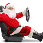 Nella foto c'è babbo natale con il suo classico vestito rosso e la barba bianca seduto su un sedile di auto con il volante retto dalla mano sinistra. Ha la cintura di sicurezza allacciata e saluta con la mano destra.
