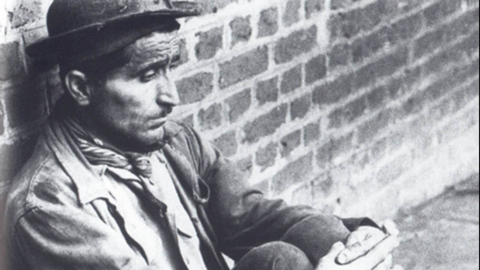 Foto in bianco e nero del minatore Rocco Romasco seduto e appoggiato ad un muro di mattoni. Indossa un cappello.