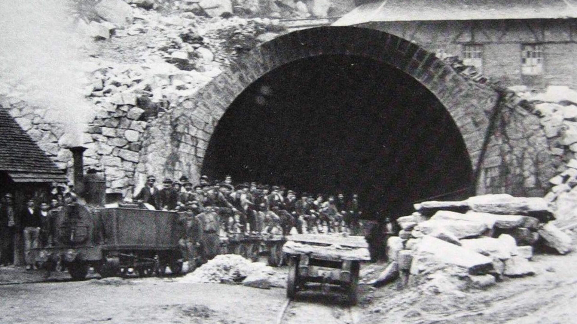 Foto in bianco e nero scattata nel 1875 all'ingresso del tunnel del Gottardo. Molti lavoratori sono stipati sui carrelli che portano in galleria